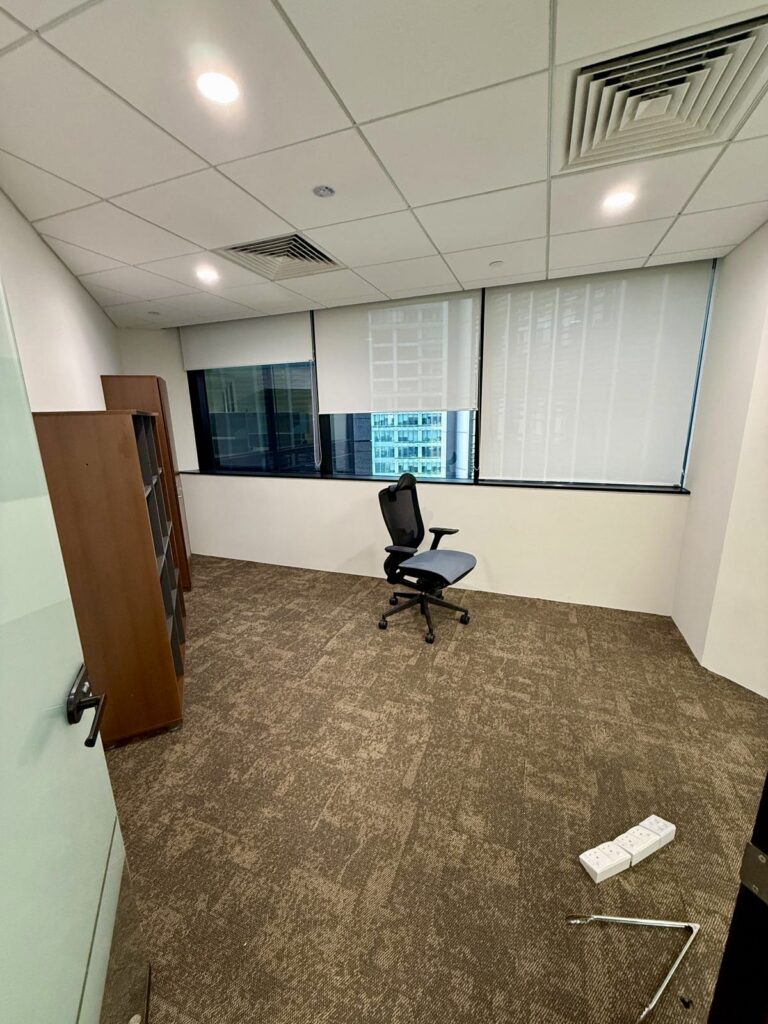 Empty office room with brown carpet, a black office chair, a wooden bookcase, large windows with blinds, and a power strip on the floor.