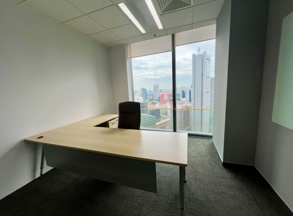 A modern, empty office with a large window showing a city skyline, a single desk, an office chair, and grey carpeted flooring.