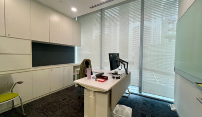 Modern office with white cupboards, a desk with computer monitor, chair, water bottle, and snacks. Large windows with blinds show city buildings outside.