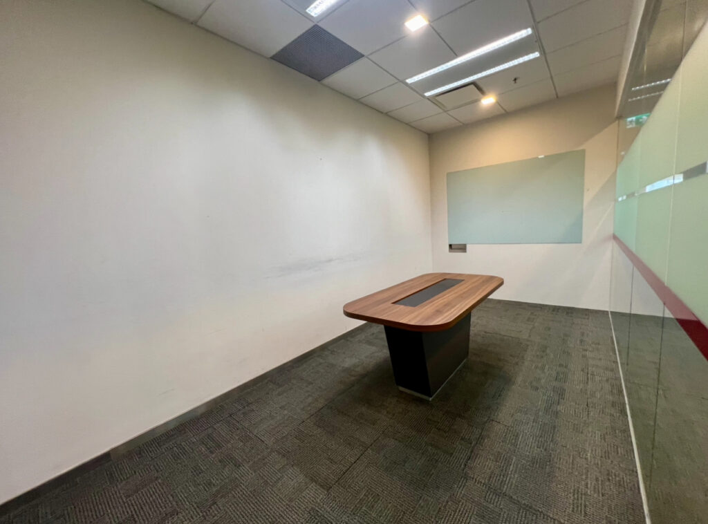 A small, empty meeting room with a single rectangular table, a glass wall on one side, carpeted floor, and a whiteboard on the wall.