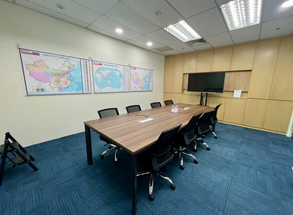 A conference room with a wooden table, six black chairs, wall maps, a flat-screen TV, and bottled water on the table, featuring blue carpet and overhead lights.