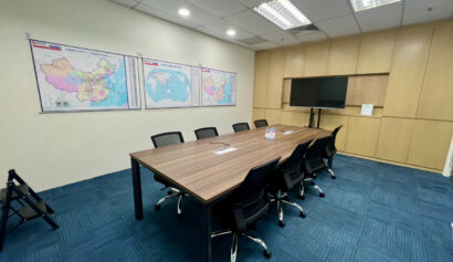 A conference room with a wooden table, six black chairs, wall maps, a flat-screen TV, and bottled water on the table, featuring blue carpet and overhead lights.