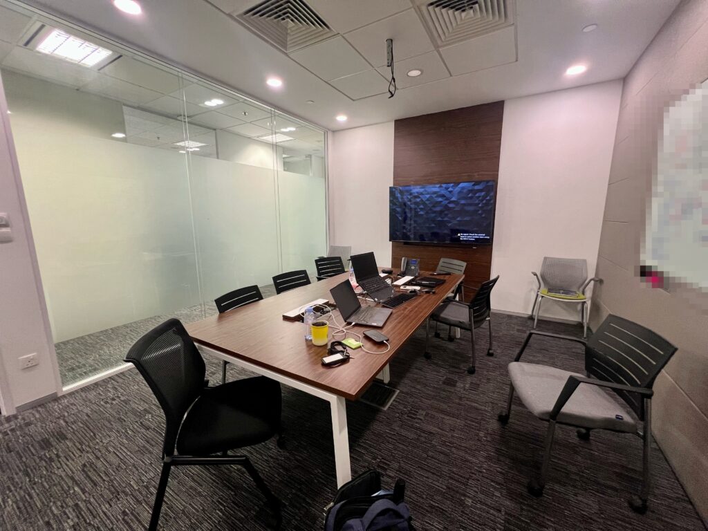 A modern conference room with a wooden table, office chairs, laptops, and a wall-mounted screen. Glass walls separate the room from the office outside.