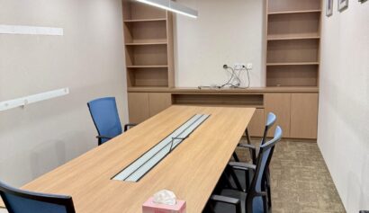 A modern conference room with a wooden table, five blue office chairs, empty shelves, a box of tissues, wall pictures, and overhead lighting.