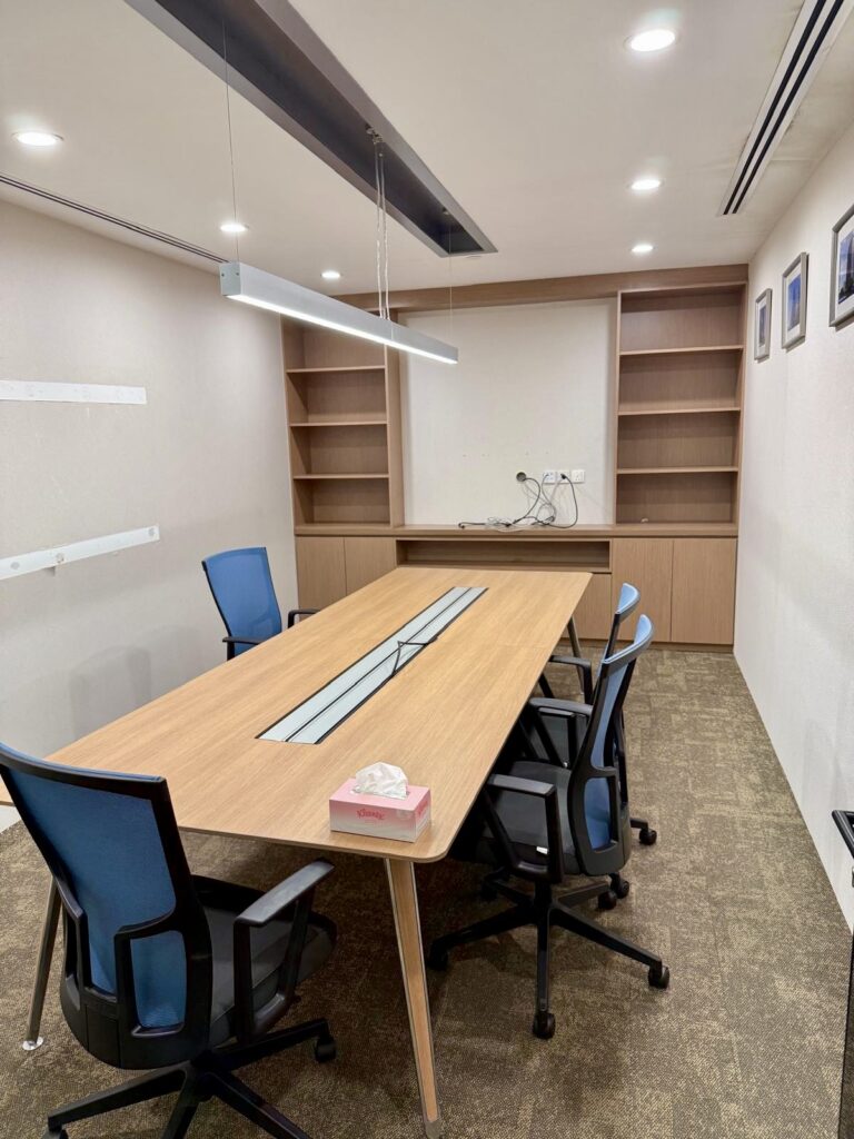 A modern conference room with a wooden table, five blue office chairs, empty shelves, a box of tissues, wall pictures, and overhead lighting.