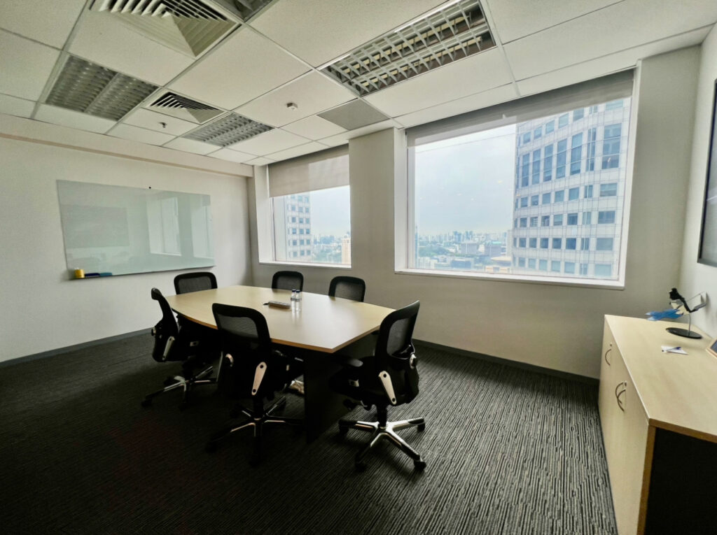 A modern conference room with six chairs around a table, two large windows, a whiteboard, and a cupboard with a lamp and stationery.
