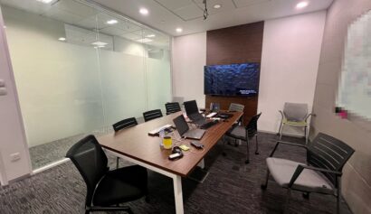 A modern conference room with a wooden table, office chairs, laptops, and a wall-mounted screen. Glass walls separate the room from the office outside.