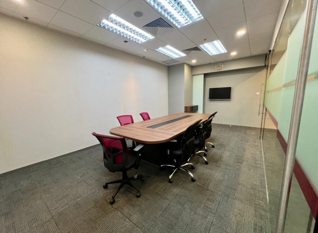 A modern conference room with a long wooden table, eight office chairs, a wall-mounted TV, and glass walls on one side.