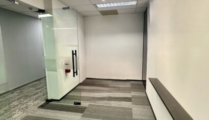 Empty corridor with glass door on the left, grey striped carpet, white walls, and an illuminated exit sign on the ceiling.