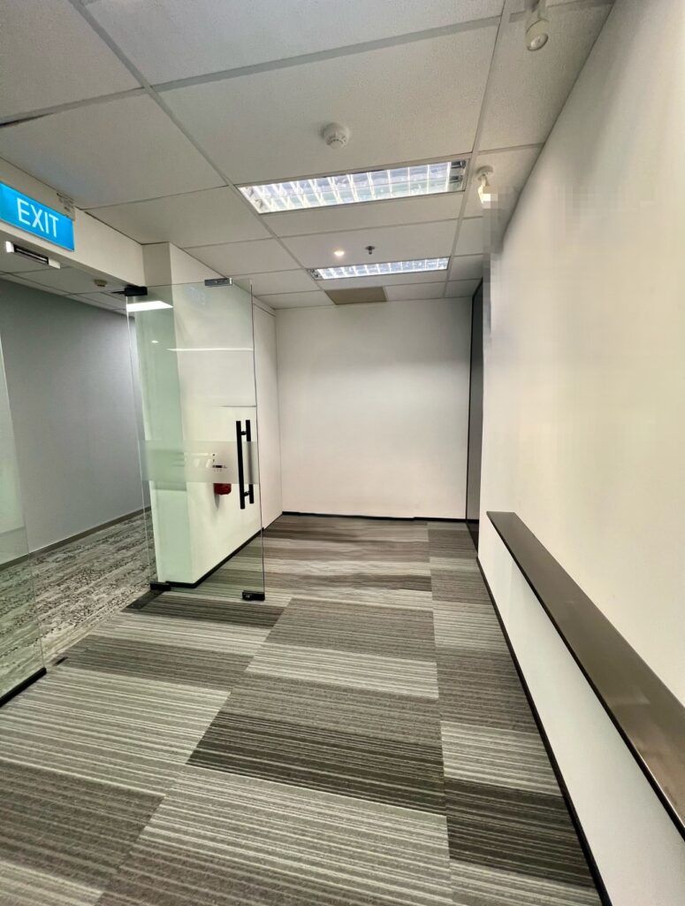Empty corridor with glass door on the left, grey striped carpet, white walls, and an illuminated exit sign on the ceiling.