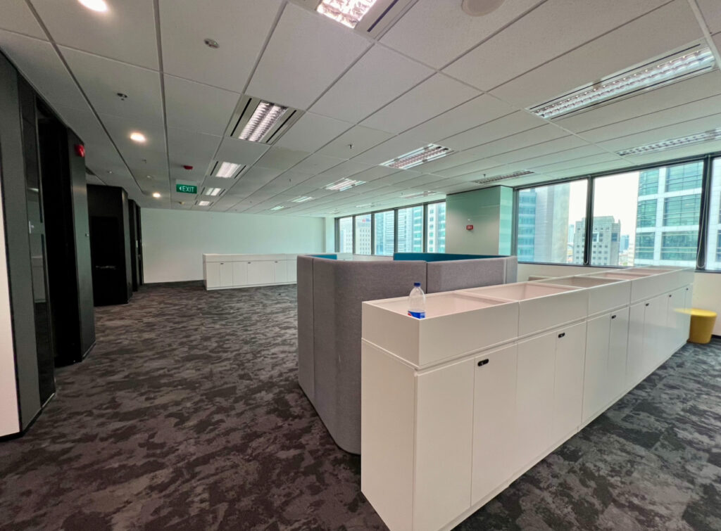 Modern office space with grey carpet, white storage cupboards, a grey partition, large windows, and natural light. A water bottle is on one cupboard and city buildings are visible outside.