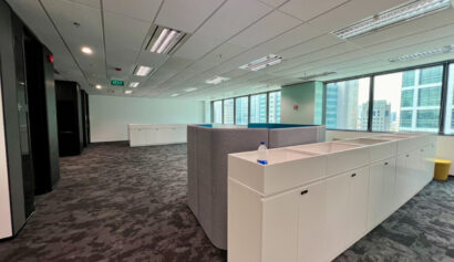 Modern office space with grey carpet, white storage cupboards, a grey partition, large windows, and natural light. A water bottle is on one cupboard and city buildings are visible outside.