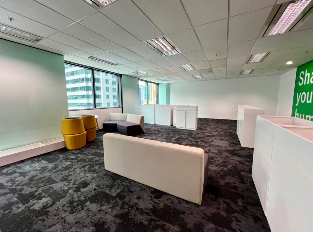 Modern office space with grey carpet, beige and black sofas, yellow chairs, white desks, and large windows overlooking city buildings.