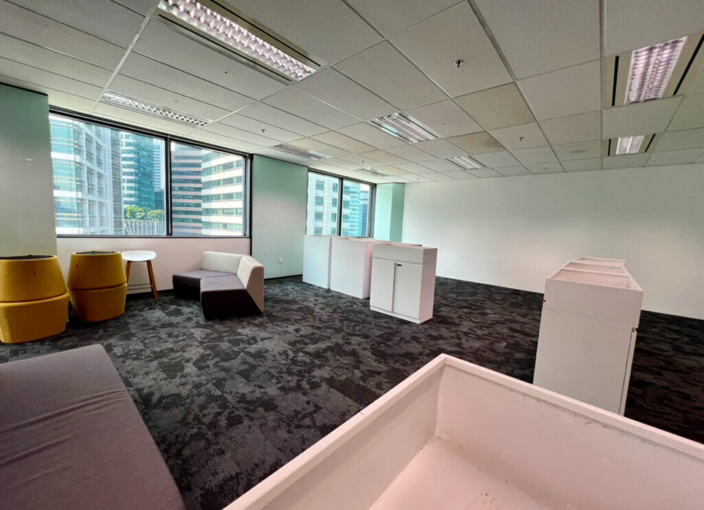 Modern office space with large windows, a few chairs, sofas, tables, and cupboards on a dark carpeted floor. The room appears spacious and minimally furnished.