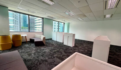 Modern office space with large windows, a few chairs, sofas, tables, and cupboards on a dark carpeted floor. The room appears spacious and minimally furnished.
