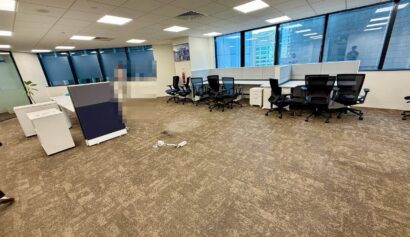 Spacious modern office with multiple empty desks and chairs, large windows, brown carpet, and a few scattered office items on the floor.