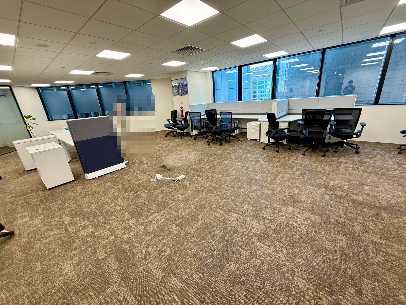 Spacious modern office with multiple empty desks and chairs, large windows, brown carpet, and a few scattered office items on the floor.
