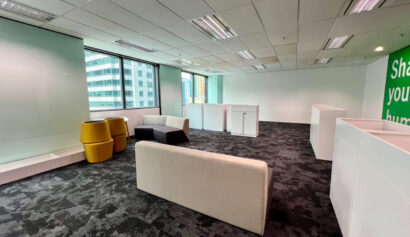 Modern office space with grey carpet, beige and black sofas, yellow chairs, white desks, and large windows overlooking city buildings.