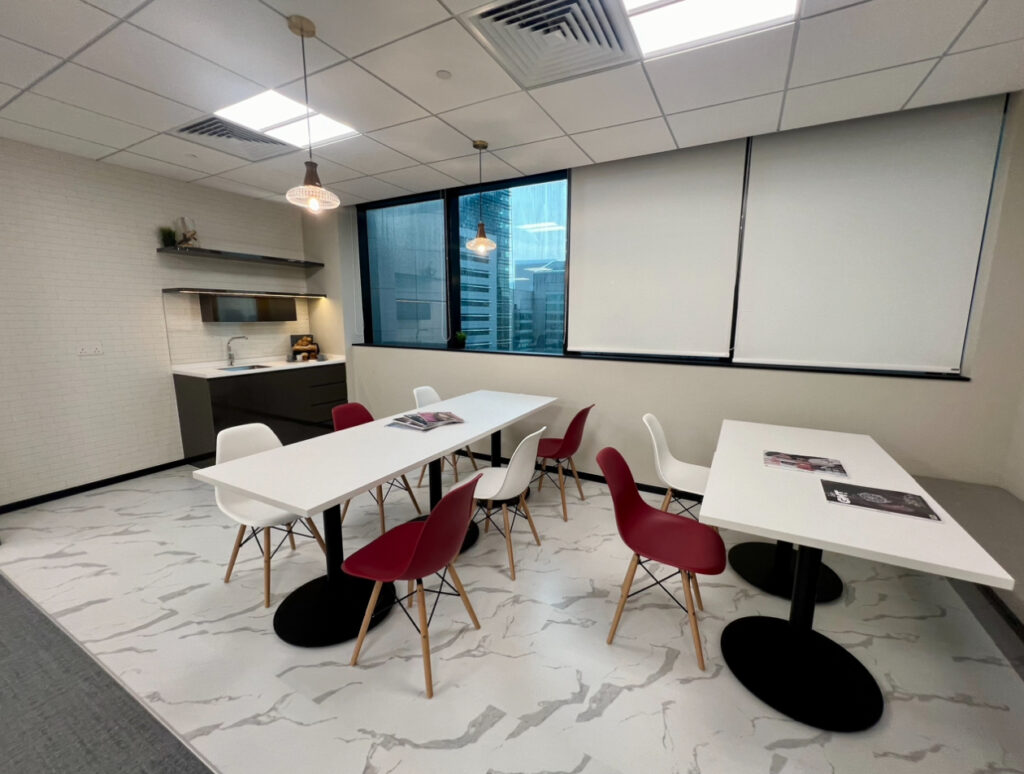 Modern office break room with white tables, red and white chairs, a kitchenette, and large windows with blinds, featuring a marble-patterned floor and overhead lighting.