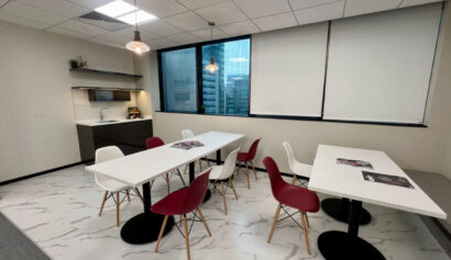 Modern office break room with white tables, red and white chairs, a kitchenette, and large windows with blinds, featuring a marble-patterned floor and overhead lighting.