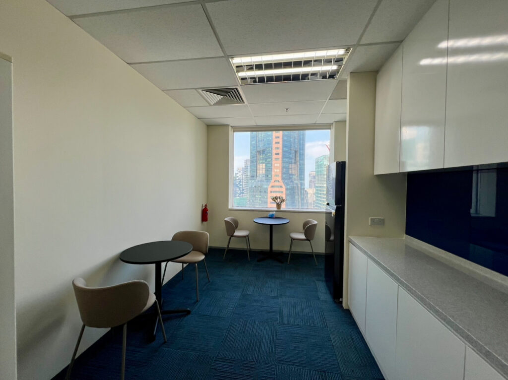 Modern office kitchenette with blue carpet, white cupboards, two round tables, four chairs, and a window overlooking city buildings. A small plant is placed on the windowsill.