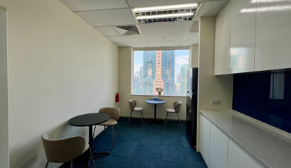 Modern office kitchenette with blue carpet, white cupboards, two round tables, four chairs, and a window overlooking city buildings. A small plant is placed on the windowsill.