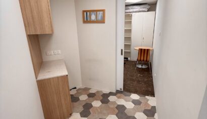 Narrow room with hexagonal tile flooring, a small worktop with wooden cupboards, a sliding door, wall art, and a view into an adjacent office space with a round table and shelves.