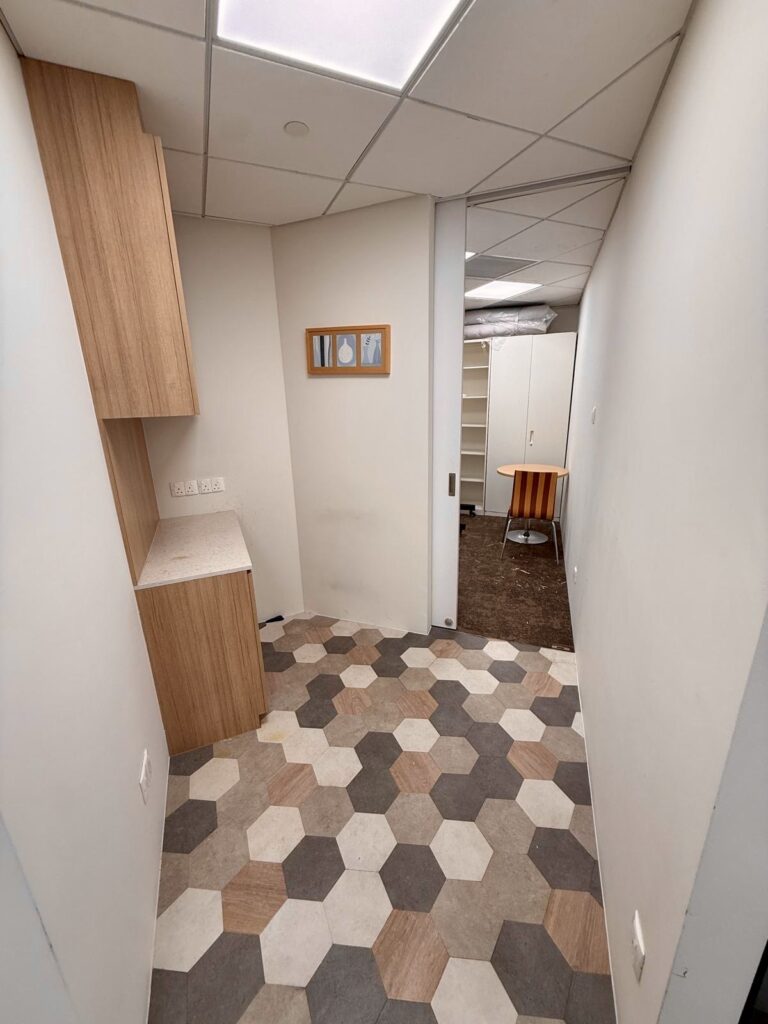 Narrow room with hexagonal tile flooring, a small worktop with wooden cupboards, a sliding door, wall art, and a view into an adjacent office space with a round table and shelves.