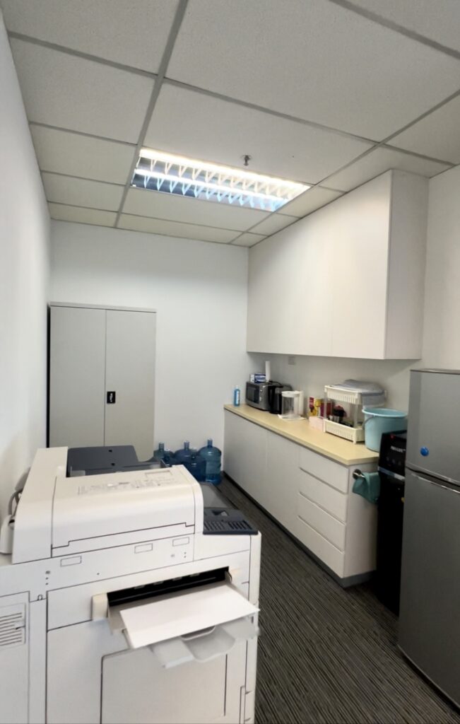 Office break room with a printer, water dispensers, countertop appliances, cupboards, and a fridge under fluorescent ceiling lights.