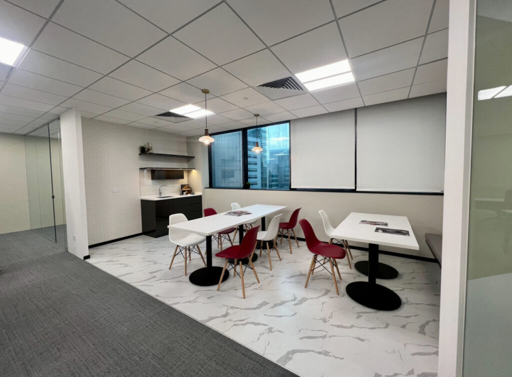 Modern office kitchenette and dining area with white tables, red and white chairs, pendant lights, and large windows with blinds. A kitchenette counter with shelves is in the corner.