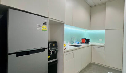A modern office pantry with a fridge, microwave, sink, countertop appliances, white cupboards, and a bin on a grey floor.