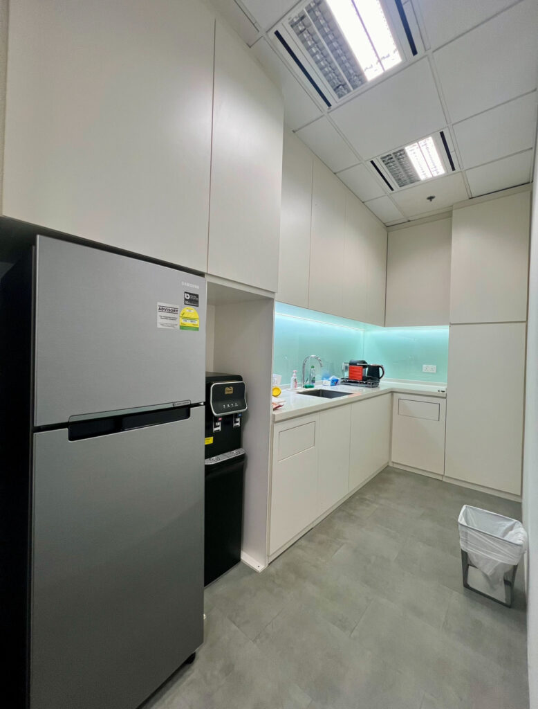 A modern office pantry with a fridge, microwave, sink, countertop appliances, white cupboards, and a bin on a grey floor.