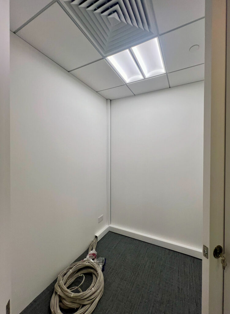 A small empty room with white walls, grey carpet, ceiling lights, a vent, and a coiled bundle of cables on the floor near one wall.