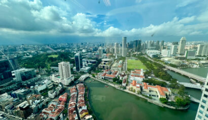 Aerial view of a city with a river running through it, high-rise buildings, a park, and densely-packed houses under a partly cloudy sky.