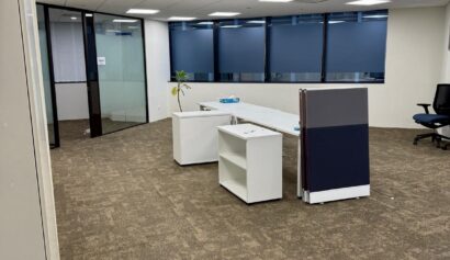 An empty office space with brown carpet, white desks, partition panels, a potted plant, swivel chairs, and large windows with blue blinds.
