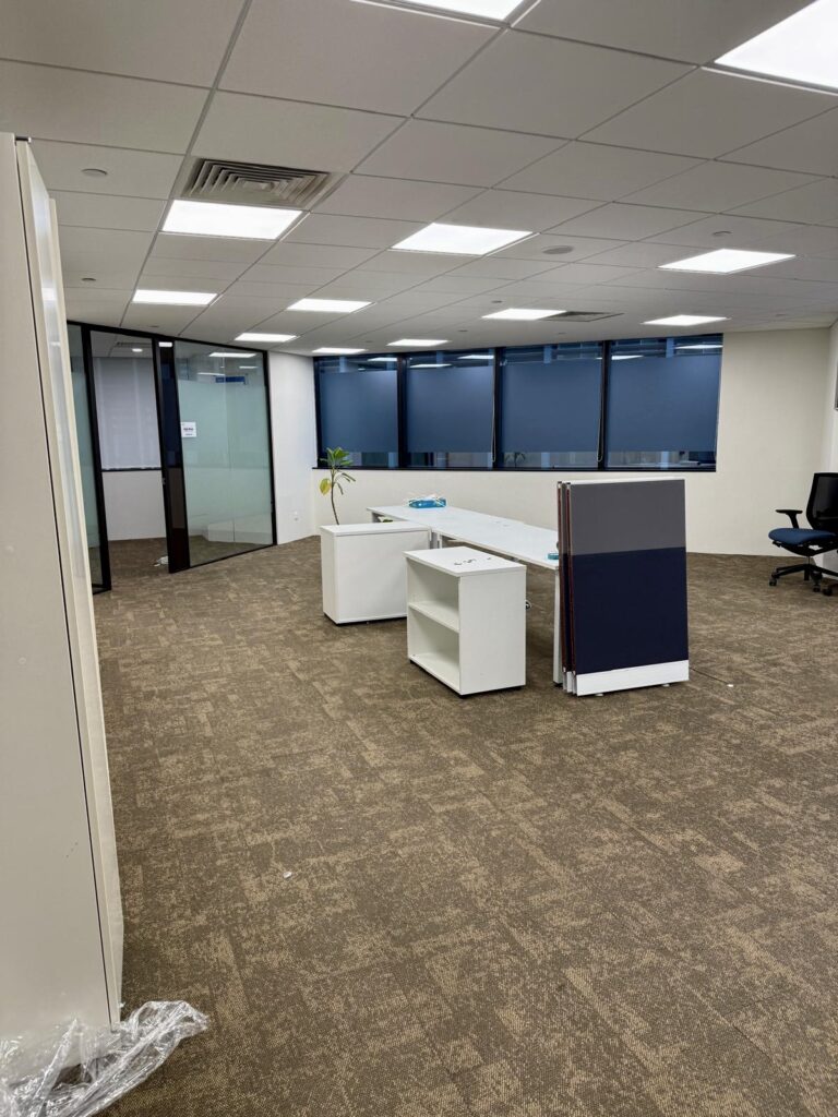 An empty office space with brown carpet, white desks, partition panels, a potted plant, swivel chairs, and large windows with blue blinds.