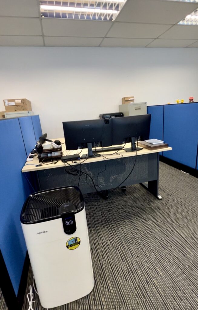 A modern office workspace with two monitors on a desk, a headset, office supplies, and a white air purifier positioned in front of the desk. Blue cupboards line the background.
