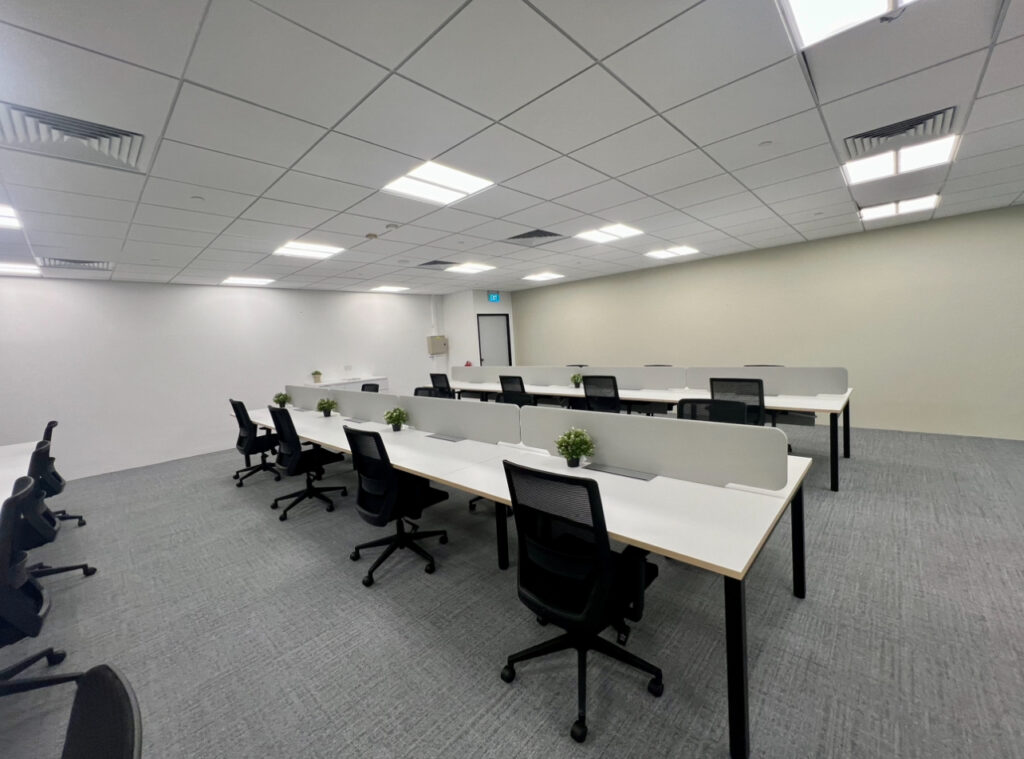 A modern, empty office space with white desks, black chairs, partition dividers, and small potted plants on each desk under bright ceiling lights.
