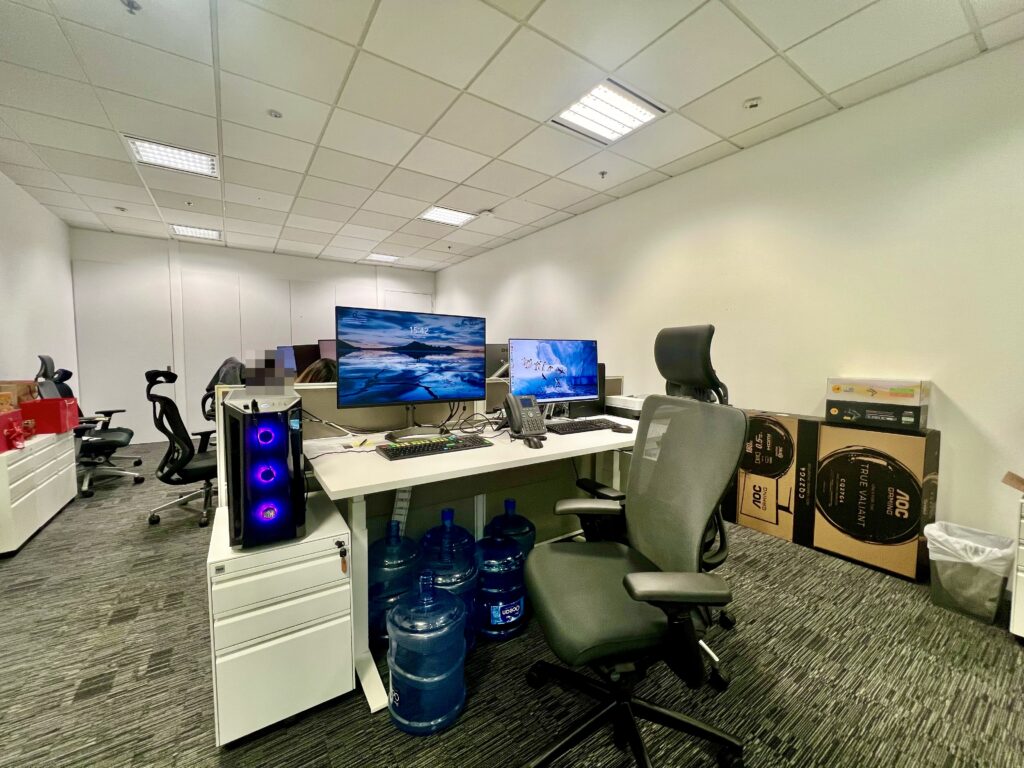 Modern office workspace with dual monitors, office chairs, a desktop computer, water jugs under the desk, and cardboard boxes stacked in the corner.