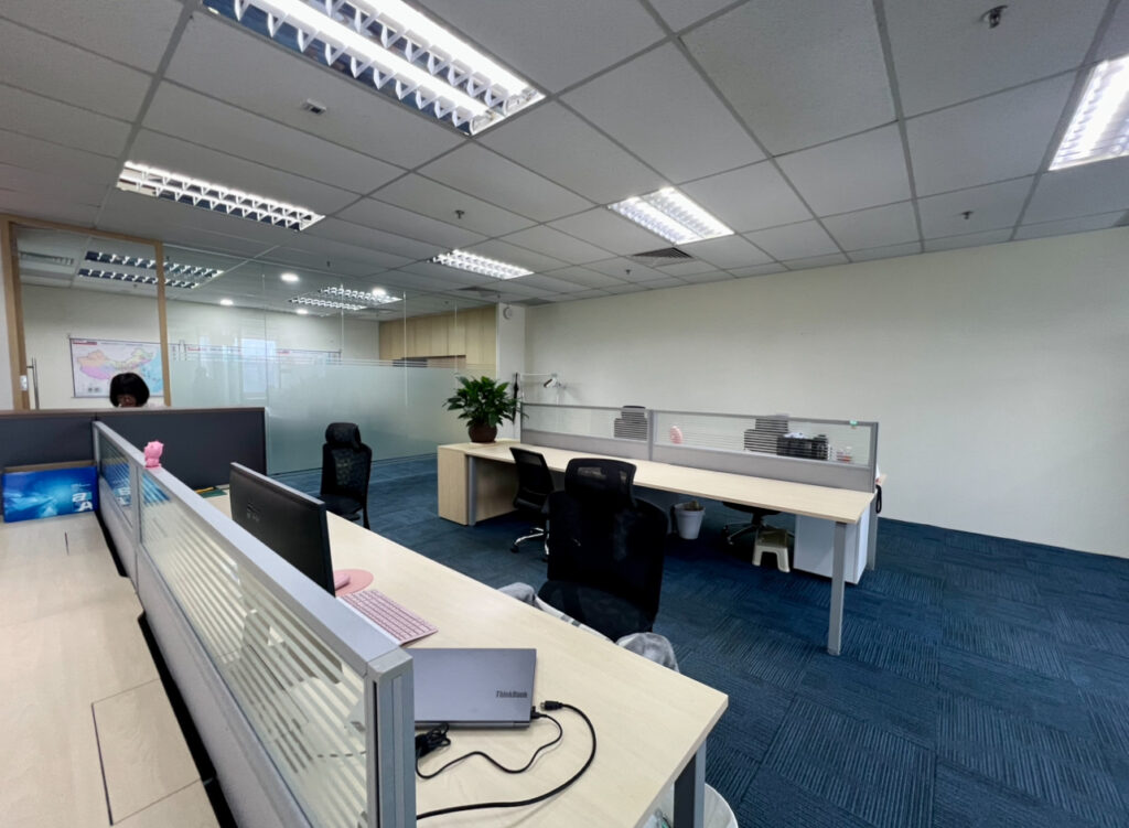 Modern open-plan office with several empty desks, office chairs, computers, partition screens, and blue carpeted flooring under fluorescent lighting.