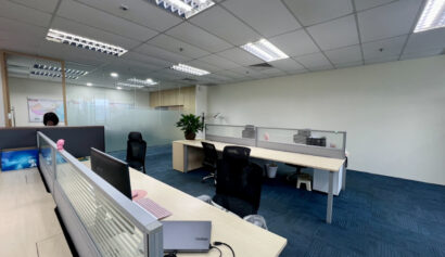 Modern open-plan office with several empty desks, office chairs, computers, partition screens, and blue carpeted flooring under fluorescent lighting.