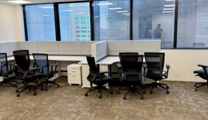 Empty office space with several black chairs, white desks, and large windows overlooking modern buildings.