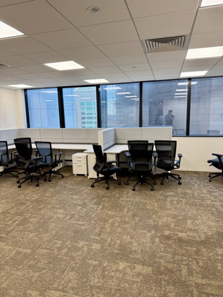 Empty office space with several black chairs, white desks, and large windows overlooking modern buildings.