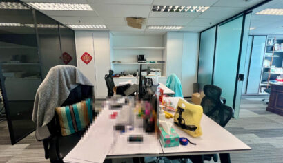 Modern office with desks, swivel chairs, assorted items and clutter on the tables; shelves and cupboards in the background; ceiling tiles partially removed.