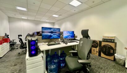 Modern office workspace with dual monitors, office chairs, a desktop computer, water jugs under the desk, and cardboard boxes stacked in the corner.
