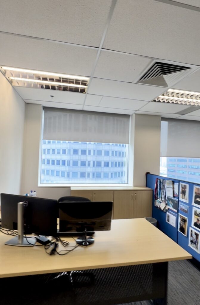 Office workspace with two computer monitors on a desk, a window with blinds, a cupboard, and a partition with various photos and lanyards displayed.