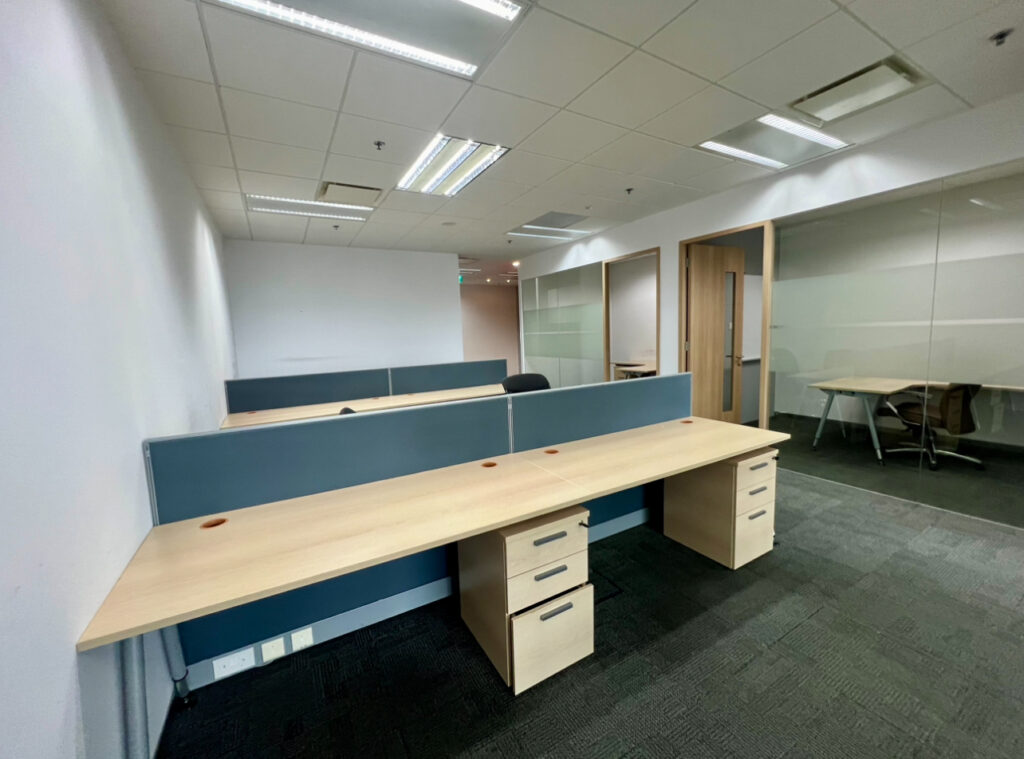 Empty modern office space with several wooden desks, partition dividers, swivel chairs, and a glass-walled meeting room in the background.