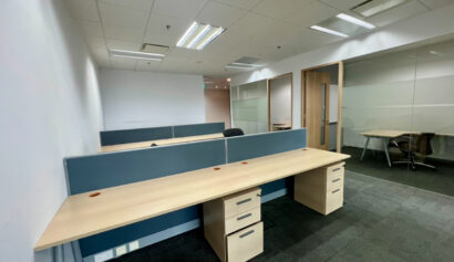 Empty modern office space with several wooden desks, partition dividers, swivel chairs, and a glass-walled meeting room in the background.