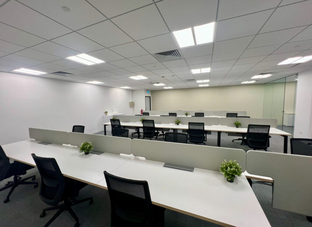 A modern open-plan office with rows of empty desks, black office chairs, partition screens, and small potted plants on the desks under bright ceiling lights.