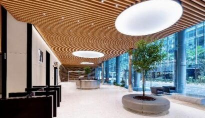 Spacious modern lobby with wooden slat ceiling, large circular lights, indoor trees, floor-to-ceiling windows, and seating areas.